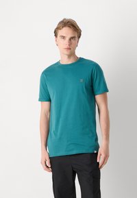 Teal majica s kratkimi rokavi iz mehke bombažne tkanine, z okroglim izrezom in majhnim oranžnim logotipom na levi strani prsnega koša. Uporabljena je bila skupaj s črnimi cargo hlačami.