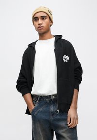 Μαύρο πλεκτό φούτερ με φερμουάρ, διακοσμημένο με λευκό λογότυπο, συνδυασμένο με απλό λευκό μπλουζάκι και ξεβαμμένο τζιν σε μπλε χρώμα. Το σύνολο ολοκληρώνεται με ένα μπεζ σκουφάκι.