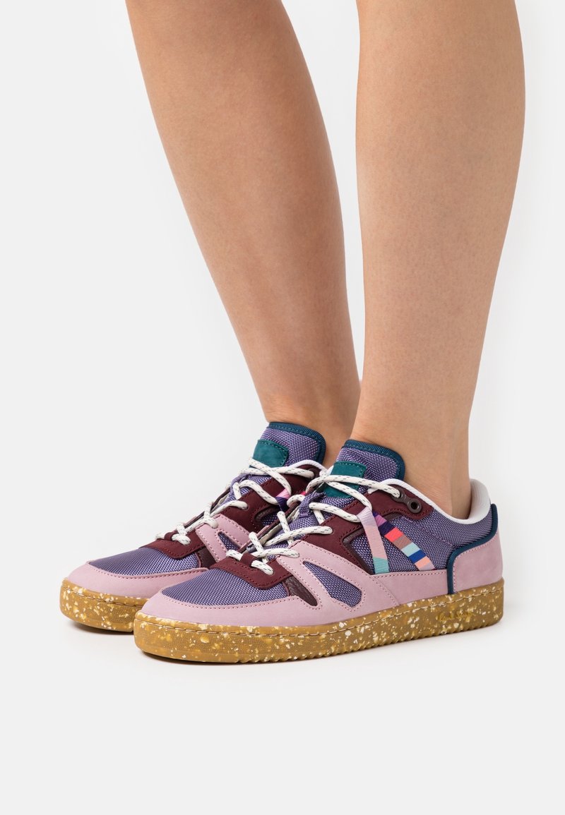 Jambes portant des baskets basses avec des panneaux violets, roses et bordeaux, des lacets blancs, des bandes latérales multicolores et des semelles en gomme tachetées sur fond blanc.