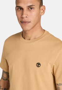 T-shirt in cotone beige con collo a giro. Presenta un piccolo logo di un albero nero nella parte sinistra del petto. Tessuto morbido, vestibilità comoda.