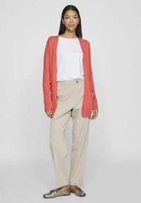 Korallenfarbener Cardigan mit gerippten Bündchen, kombiniert mit einem weißen Hemd und beigen Hosen. Die Schuhe sind metallisch und haben eine Schleifenakzent. Schlichter, lässiger Schnitt.