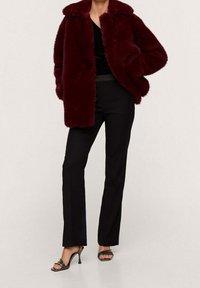 Persona vistiendo un abrigo de piel sintética burdeos, una blusa negra, pantalones rectos negros y sandalias de tacón alto negras con tiras, posando frente a un fondo liso.