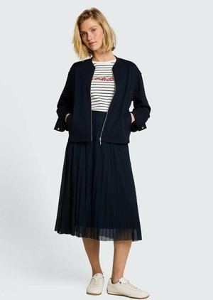 Femme portant une veste zippée noire, un t-shirt blanc rayé, une jupe midi plissée noire et des baskets blanches, debout les mains dans les poches.