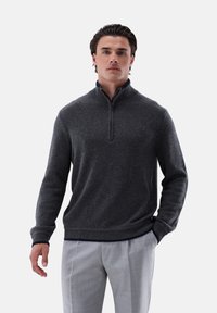 Dunkelgrauer Pullover mit Reißverschlusskragen, gerippten Bündchen und Saum. Über hellgrauen Hosen getragen, die die gestrickte Textur und Passform hervorheben.