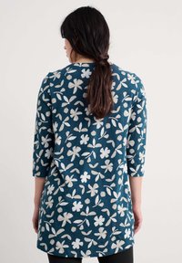 Ženska s temnimi lasmi spet zvezanimi, oblečena v temno teal tuniko z belimi cvetličnimi in listnatimi vzorci, prikazana s hrbtne strani na enobarvni podlagi.