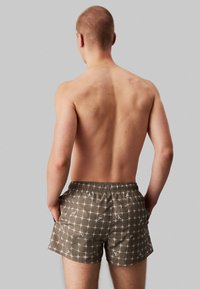 Bruine zwemshorts met een wit ruitpatroon, elastische tailleband en logo-accenten; gemaakt van lichtgewicht, snel drogend materiaal.