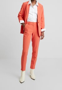 Pomarańczowy garnitur składa się z dopasowanej marynarki i spodni o wąskim kroju, zestawiony z białą koszulą w paski. Modelka nosi kremowe botki za kostkę.