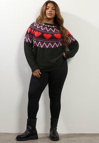 Pull en tricot noir avec motif de cœur rouge, accents géométriques rose et blanc, associé à des leggings noirs et des bottines noires.