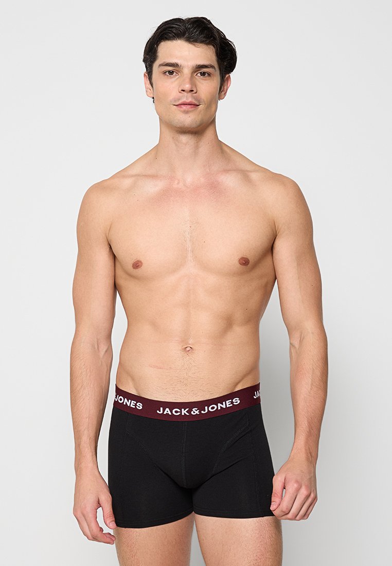 Hombre sin camiseta con cabello oscuro vistiendo calzoncillos negros de Jack & Jones, de pie contra un fondo gris claro liso.