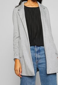 Mujer vistiendo un blazer de rayas grises sobre una blusa negra plisada y jeans azules, de pie frente a un fondo gris liso.