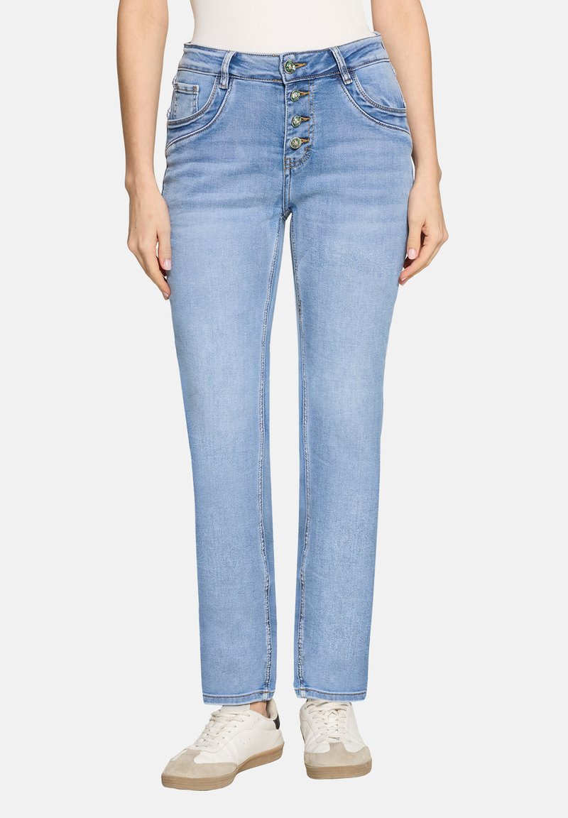 Kvinde iført lyseblå straight-leg jeans med fire synlige knapper og hvide sneakers, stående mod en ensfarvet baggrund.