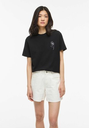 Jeune femme aux cheveux foncés mi-longs, portant un t-shirt noir avec un motif de fleur blanche et un short blanc, debout devant un fond uni.