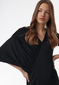 Vestido negro con escote en V, que presenta fruncidos laterales y mangas anchas. Está hecho de una tela suave con un corte holgado.