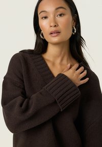 Pull en tricot marron avec un col en V et des poignets côtelés. Le matériau est texturé, offrant une coupe ample qui met l'accent sur le confort.
