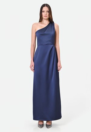 Donna che indossa un abito in raso blu navy senza maniche, lungo fino al pavimento, con una spalla e tacchi alti metallici, in piedi su uno sfondo bianco.