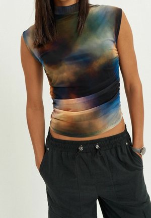 Kobieta ubrana w bezrękawnik z wysokim kołnierzem, w wielokolorowy abstrakcyjny wzór, czarne spodnie ze ściągaczem i zegarek na nadgarstku, z rękami w kieszeniach.