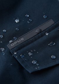 Marine blauwe stof met waterdruppels, featuring een zwart etiket met de opdruk "OPUS." Het oppervlak heeft een gladde textuur en een matte afwerking.