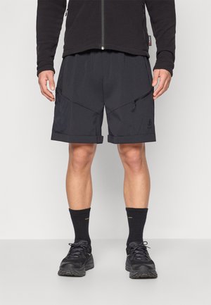 Mann trägt schwarze sportliche Shorts mit Reißverschlusstasche, schwarze Socken und schwarze Turnschuhe, steht vor einem einfarbigen Hintergrund.