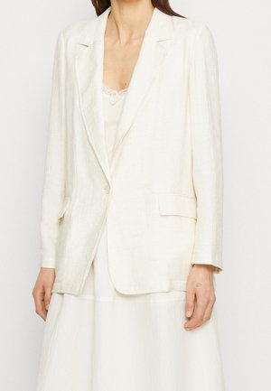 Femme portant un blazer texturé blanc cassé sur un haut orné de dentelle et une jupe assortie, debout devant un fond uni.