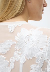 Ξανθιά γυναίκα γυρισμένη πλευρικά, που φοράει διάφανο λευκό τοπ με μεγάλο υφασμάτινο κέντημα σε μορφή λουλουδιών στην πλάτη.