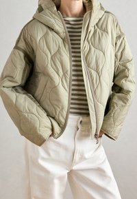 Veste matelassée vert clair avec capuche, dotée d'une fermeture éclair et d'une texture ondulée, portée par-dessus une chemise rayée, associée à un jean blanc.