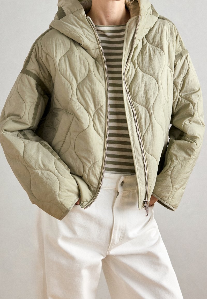 Veste matelassée vert clair avec capuche, dotée d'une fermeture éclair et d'une texture ondulée, portée par-dessus une chemise rayée, associée à un jean blanc.