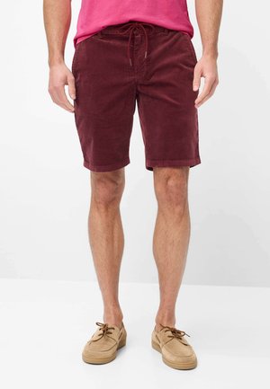Mann trägt burgunderfarbene Cordshorts mit Kordelzug und beige Schnürschuhe, steht vor einem einfarbigen Hintergrund.