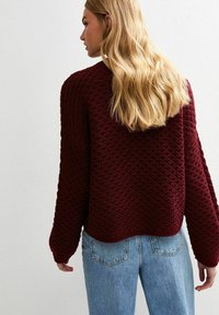 Blonde vrouw in een gestructureerde bordeauxrode trui en lichtblauwe spijkerbroek, met de rug naar de camera toe tegen een effen lichte achtergrond.