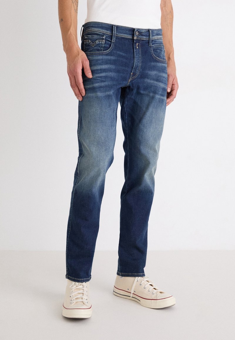 Homme portant un jean bleu délavé et des baskets en toile blanche, debout devant un fond blanc uni.