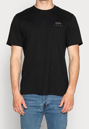 Mies, jolla on musta lyhythihainen T-paita, jossa vasemmalla rinnalla teksti "EDWIN" ja japanilaisia merkkejä, yhdistettynä sinisiin farkkuihin.