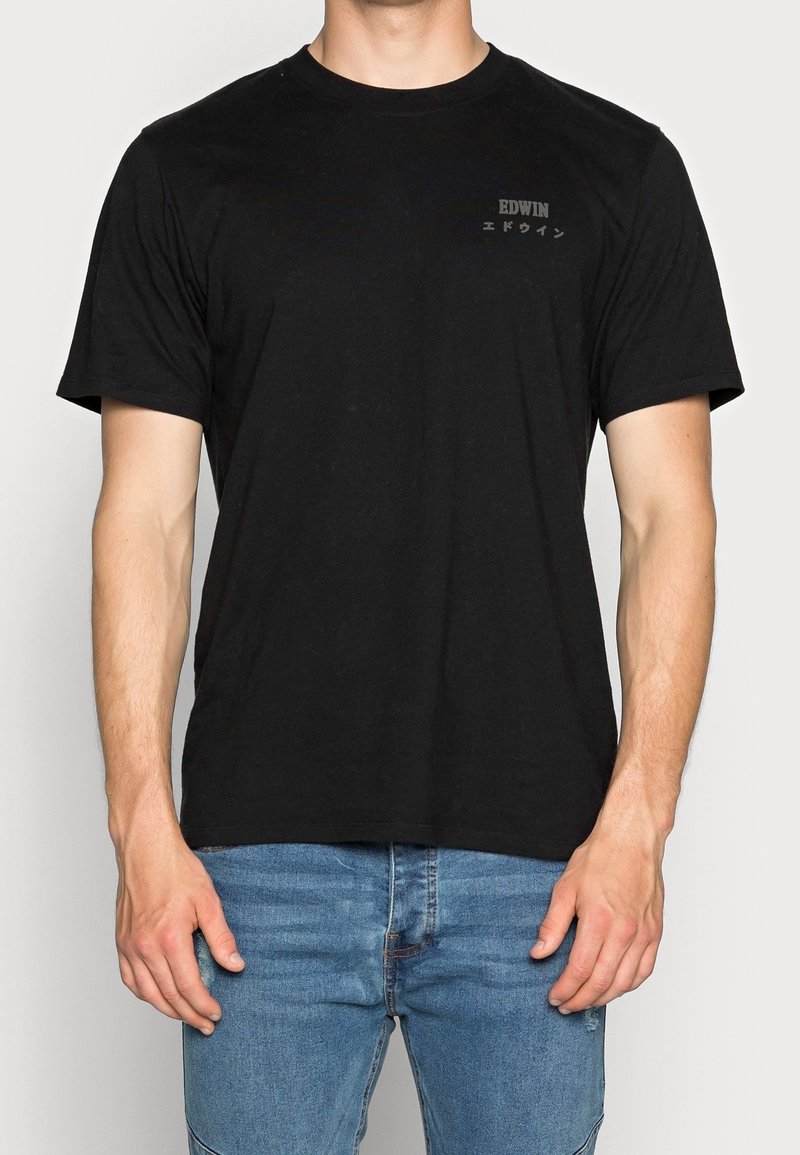 Mężczyzna w czarnej koszulce z krótkim rękawem z napisem "EDWIN" i japońskimi znakami po lewej stronie klatki piersiowej, w połączeniu z niebieskimi dżinsami.