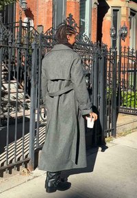 Szary wełniany płaszcz trench z zawiązywanym paskiem, długie rękawy i czarne skórzane botki do kostek, trzymający białą filiżankę, na tle żelaznej bramy.