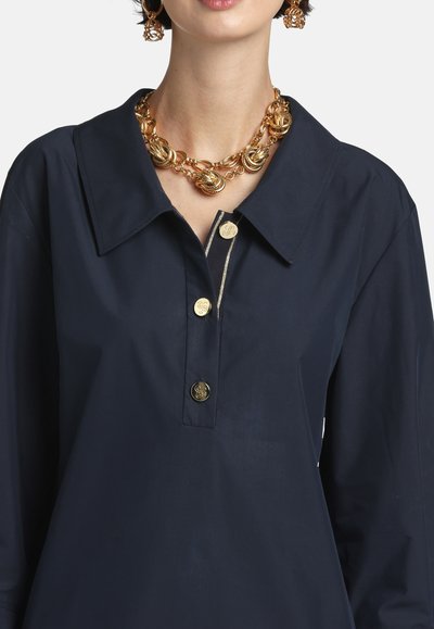 Žena v tmavomodrej košeli so zlatými gombíkmi, hrubým zlatým reťazovým náhrdelníkom a zlatými kruhovými náušnicami, ktoré sa k tomu hodia.