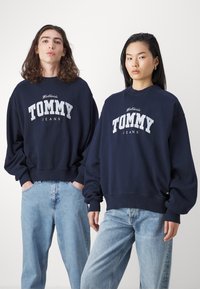 Sudaderas de gran tamaño en azul marino con el logo "TOMMY JEANS" en blanco, combinadas con jeans de un azul claro. Ajuste casual y relajado, con puños y dobladillo acanalados.