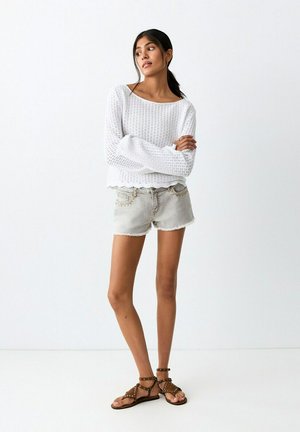 Femme vêtue d'un haut en maille blanche à manches longues, d'un short en jean clair et de sandales marron cloutées, debout contre un mur blanc uni.