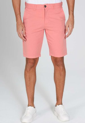 Homme portant un short corail rose jusqu'aux genoux et des baskets blanches, debout devant un fond uni.