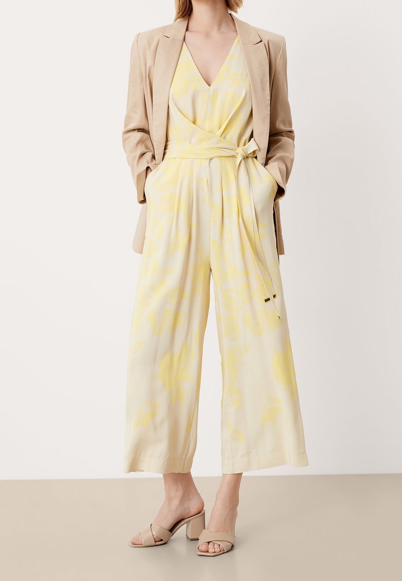 Jumpsuit floral jaune clair avec un décolleté en V, jambes larges et une ceinture à la taille, associé à un blazer beige et des sandales à talons beiges.