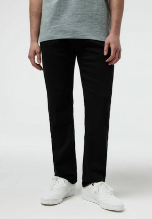 Osoba ubrana w czarne spodnie, szarą koszulę i białe buty sportowe Nike, stojąca na jednolitym jasnym tle.