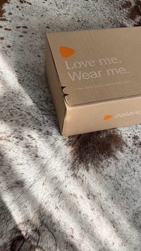 Barna kartondoboz, amelynek a tetején kivágás található, "Szeress engem. Viselj engem." felirattal és a Zalando logójával, egy mintás barna és fehér prém felületen elhelyezve.