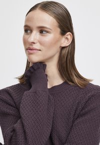 Purple textured sweater with ribbed cuffs and neckline. The fabric features a honeycomb pattern. Model poses gently touching her neck.