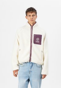 White fleece jacket with a high collar, purple zippered pocket, and embroidered design. Features a zip front and relaxed fit.