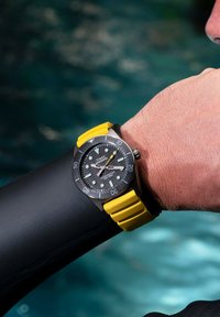Reloj de buceo negro con correa de silicona amarilla, que presenta un dial texturizado negro, marcadores de hora luminosos y un bisel de acero inoxidable.