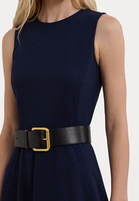 Granatowa sukienka bez rękawów z gładkiej tkaniny z zaszewkami; taliowana szerokim czarnym skórzanym paskiem z złotą metalową klamrą.