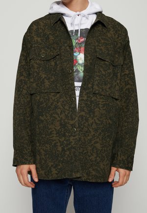Hombre con una camisa oscura verde con estampado y botones, con dos bolsillos en el pecho, sobre una sudadera blanca con diseño gráfico y vaqueros azules.