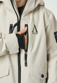 Beige weatherproof jacket with a hood, featuring black zippers, logo accents, and a textured sleeve cuff. Key designs include zippered pockets.