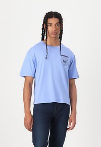 Light blue cotton T-shirt with a round neck, short sleeves, and a circular black graphic on the left chest. Worn with dark jeans.