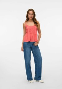 Top sin mangas de color coral con detalle de pliegues en la parte delantera, combinado con jeans de pierna ancha de color azul y zapatos blancos de punta en un fondo neutro.