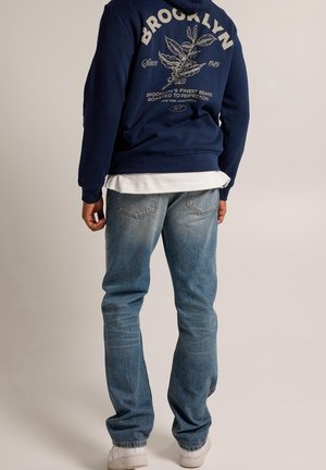 Osoba w granatowej bluzie z kapturem z napisem "BROOKLYN" i wzorem rośliny kawy na plecach, jasnoniebieskich jeansach, białej koszuli i białych trampkach, stojąca tyłem.