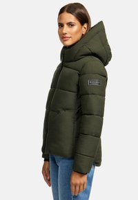 Grön pufferjacka med huva, med quiltade sektioner, framfickor och en logotyp-patch på ärmen. Bärs med blå jeans.