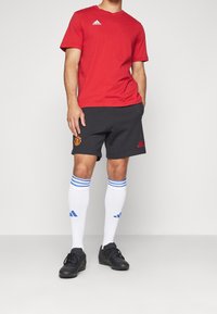T-shirt rossa a maniche corte Adidas, pantaloncini neri con logo del Manchester United, calze bianche con strisce blu e scarpe sportive nere.